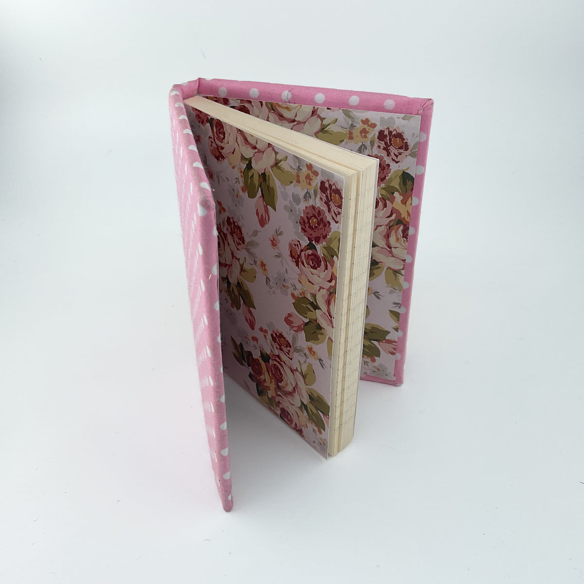 A small hardcover notebook or journal standing open on a white studio background. The cover and spine are pink with white polka dots, and the inner floral pattern features pink roses with green leaves on a pale lavender background.