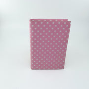 A pink notebook or journal with a white polka dot pattern, standing upright on a white background.
