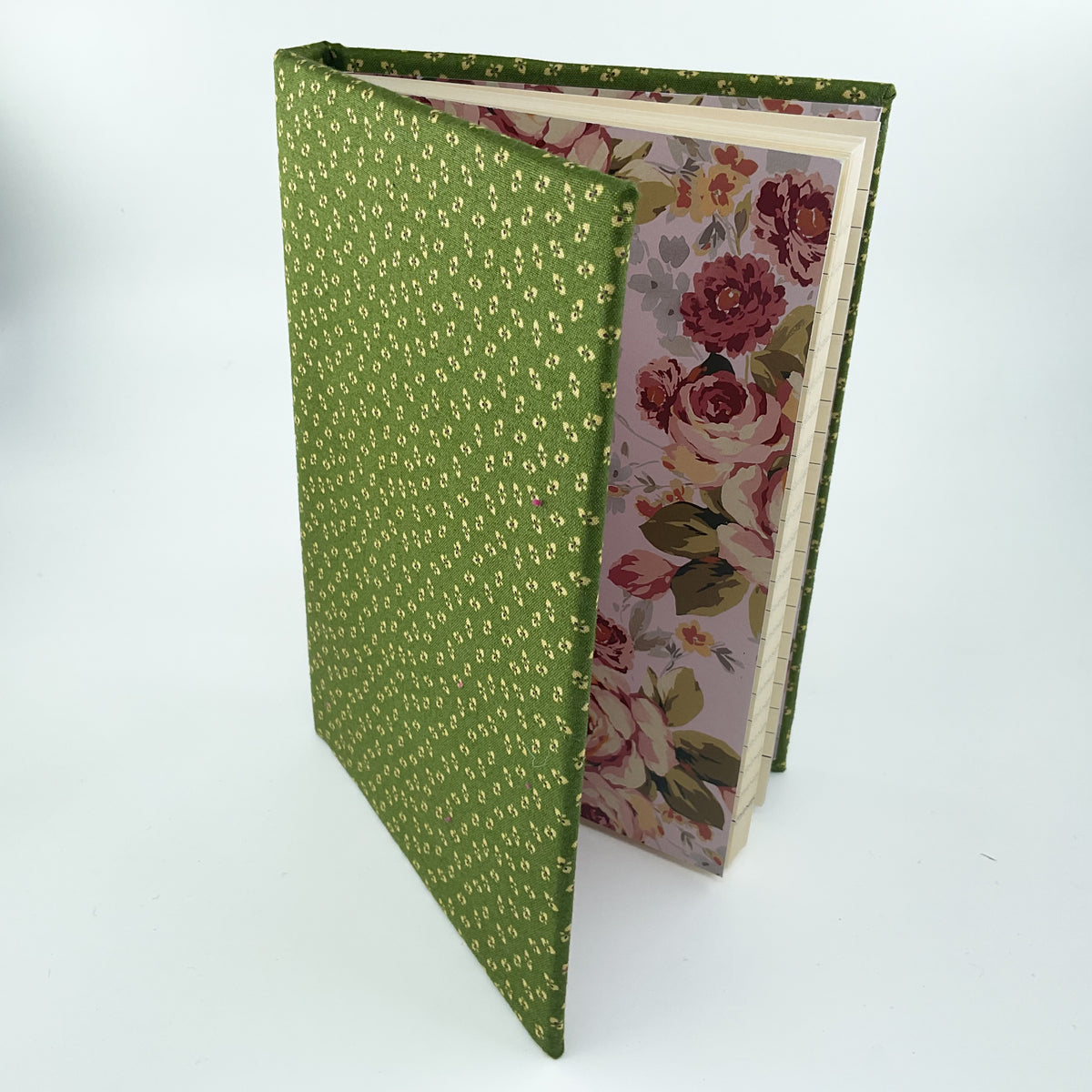 A handmade fabric-bound book or journal standing open on a white background. The cover is olive green with a small repeating gold floral pattern, and the inner pages are lined with a vintage-style floral paper featuring pink, red, and green roses.