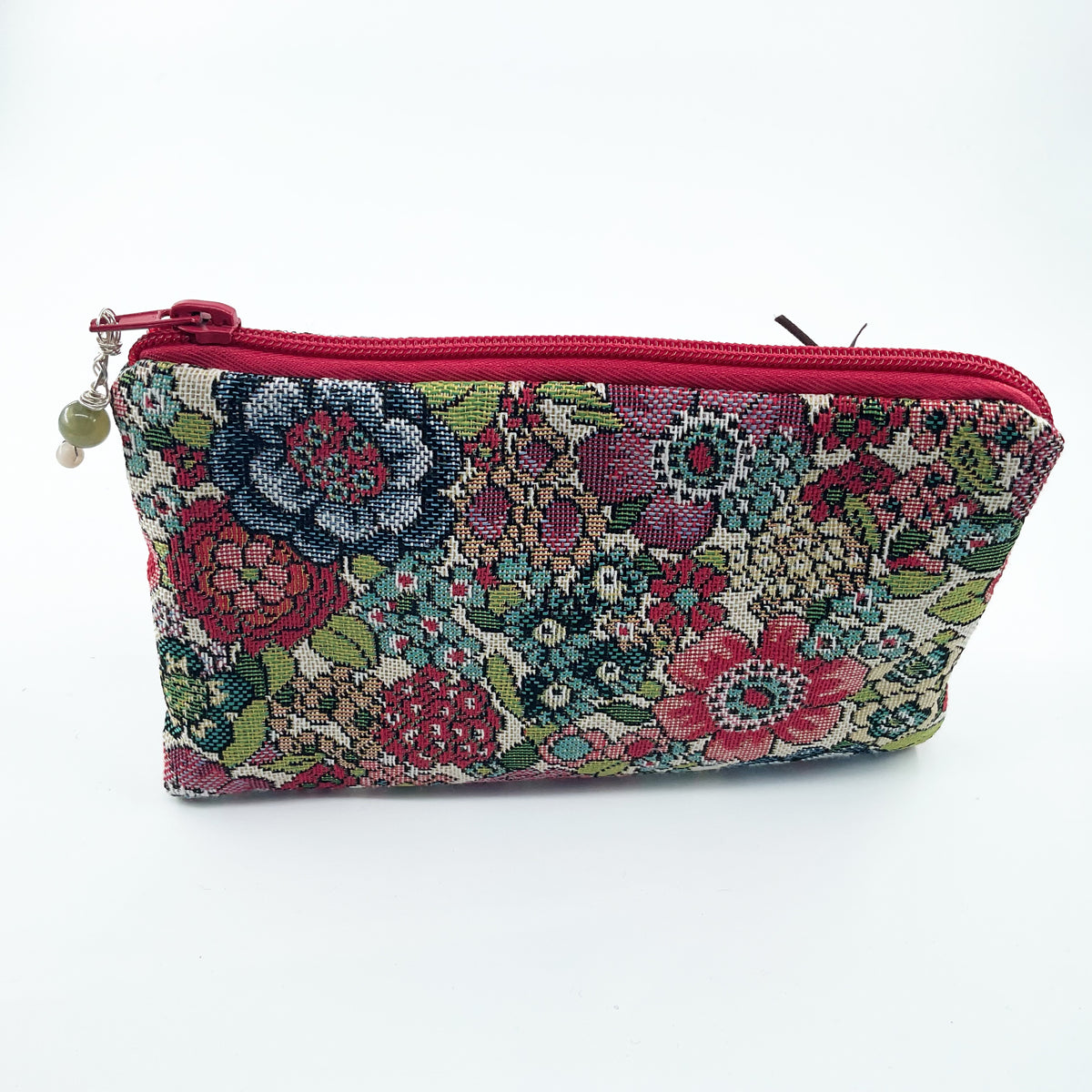 A rectangular zippered pouch with a vibrant floral tapestry pattern in red, green, blue, and beige, featuring a red zipper and a small hanging bead charm, set against a plain white background.