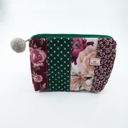 A rectangular patchwork fabric pouch with a green zipper and a gray pom-pom on the left side. The pouch features multiple vertical fabric panels in floral and patterned designs, including burgundy roses, a green patterned stripe, and peach flowers.