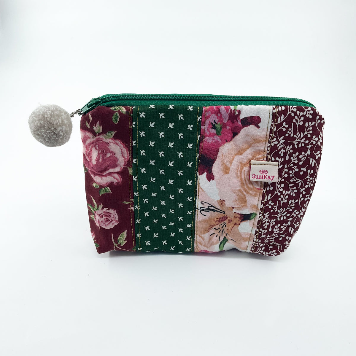 A rectangular patchwork fabric pouch with a green zipper and a gray pom-pom on the left side. The pouch features multiple vertical fabric panels in floral and patterned designs, including burgundy roses, a green patterned stripe, and peach flowers.