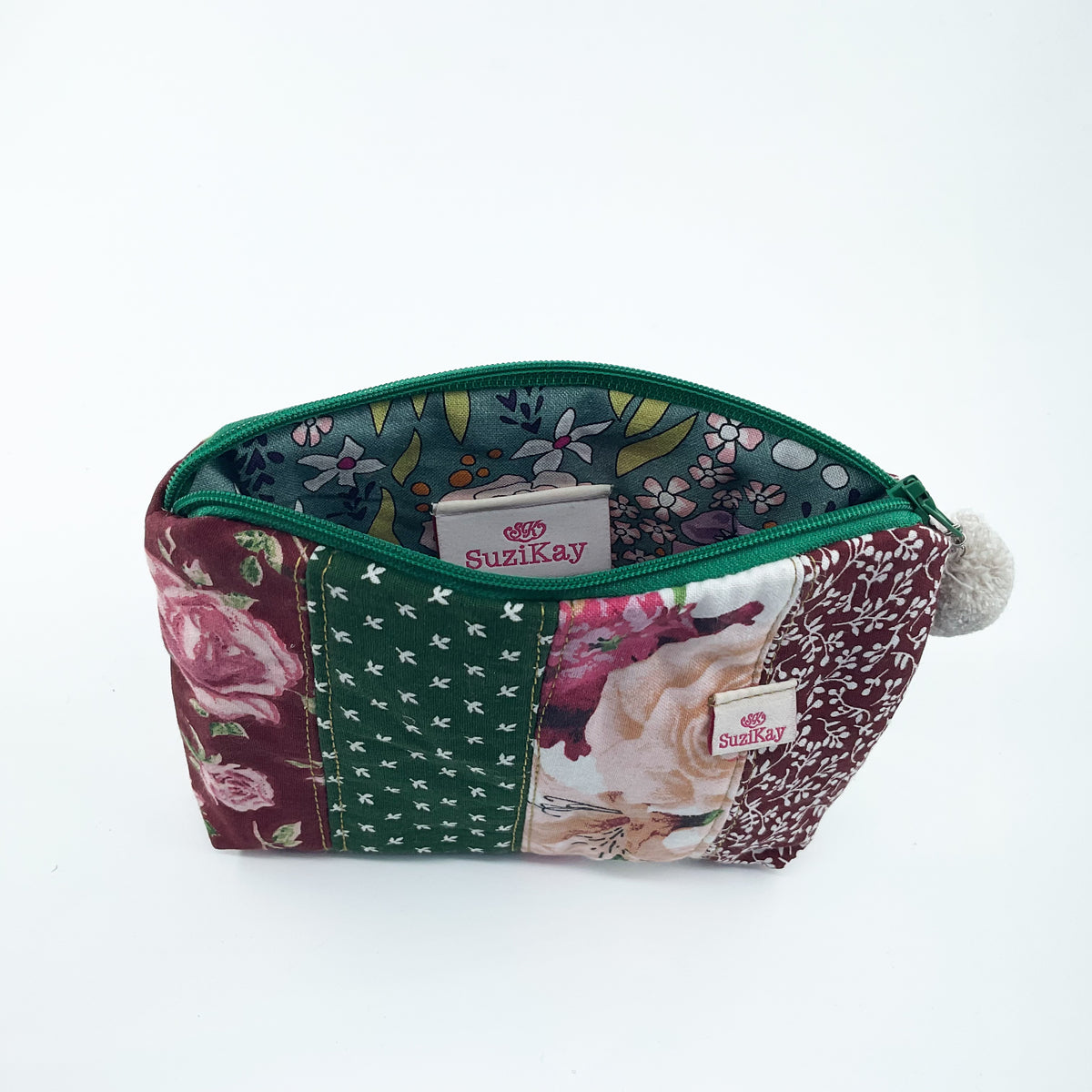 A patchwork fabric cosmetic pouch by Suzikay, featuring a green zipper, a pom-pom, and panels of floral and geometric patterns in burgundy, green, and cream, shown open on a white background.