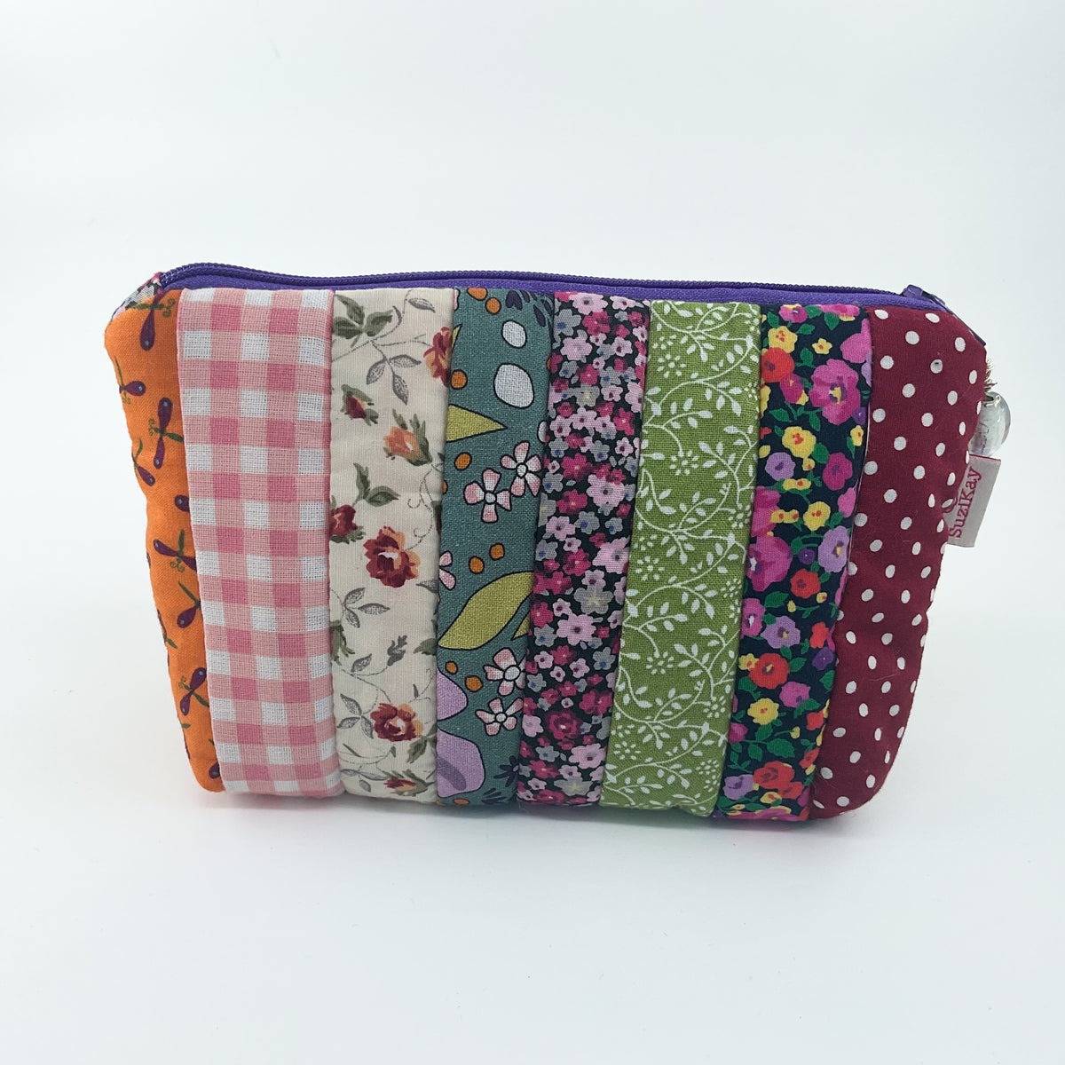 A rectangular patchwork pouch with vertical fabric panels in orange, pink gingham, cream floral, teal floral, green floral, burgundy floral, and burgundy polka dot, with a purple zipper and a small white brand tag on the right side.