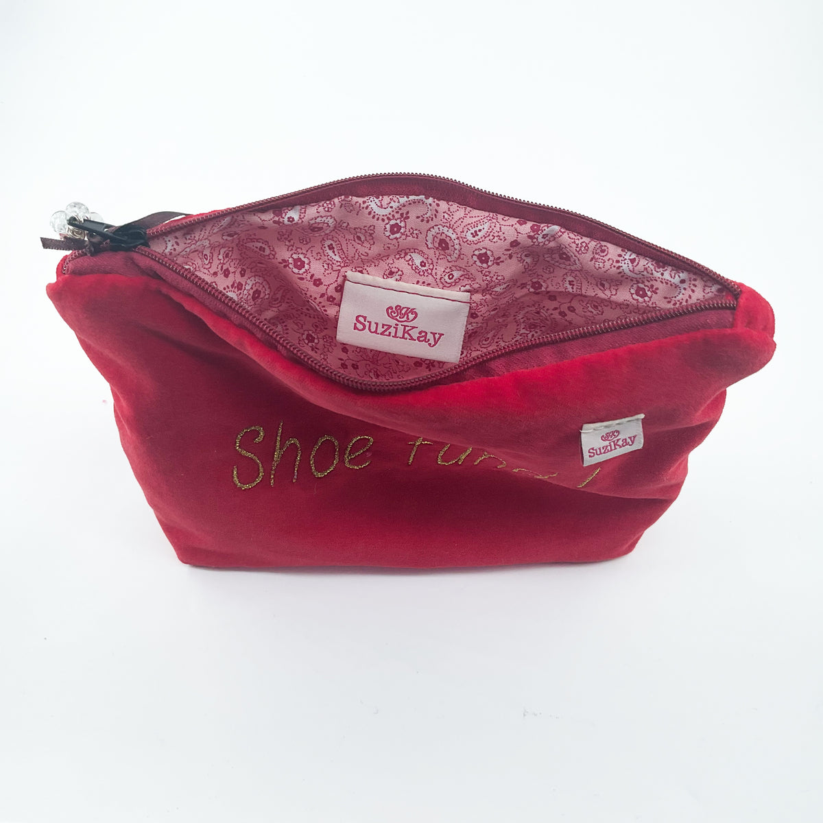 A red velvet shoe bag with a zipper closure, featuring 'Shoe Fun!' embroidered in green on the front. The interior is lined with a pink paisley pattern, and the brand label 'SuziKay' is visible inside.
