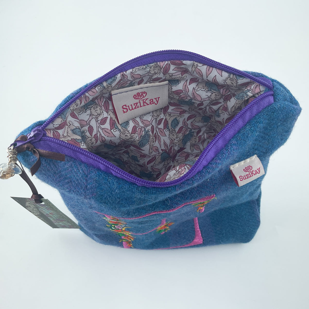 A blue denim zippered pouch with a purple zipper, featuring a floral-patterned lining and a visible 'SuziKay' brand label.
