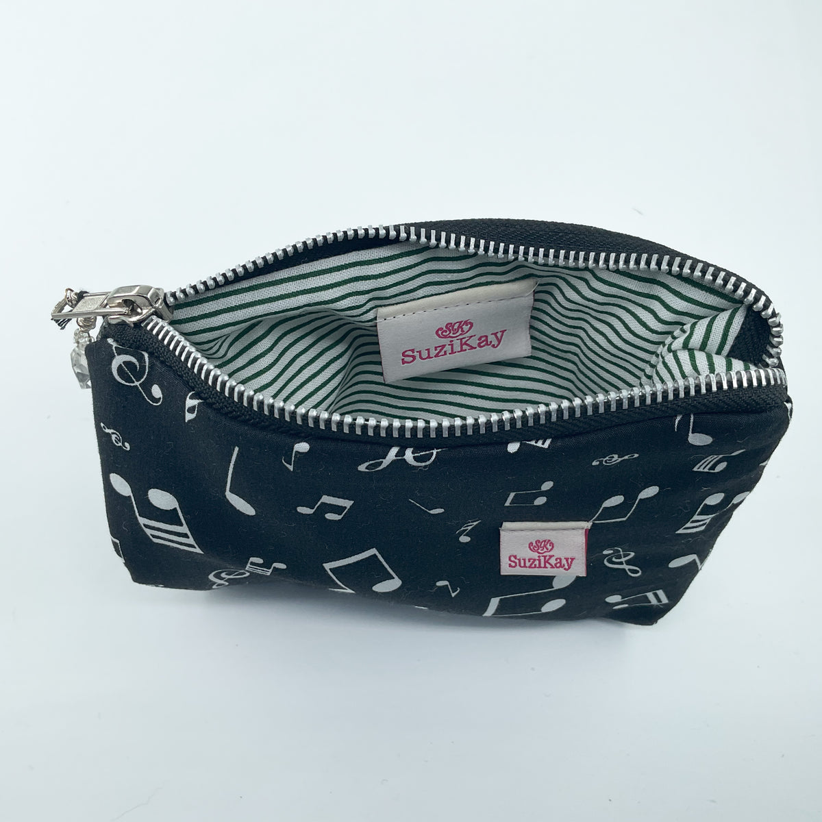 A zippered pouch with a black exterior printed with white musical notes, shown open to reveal a green and white striped interior. The brand name 'Suzikay' is visible on a label inside and on the exterior patch.