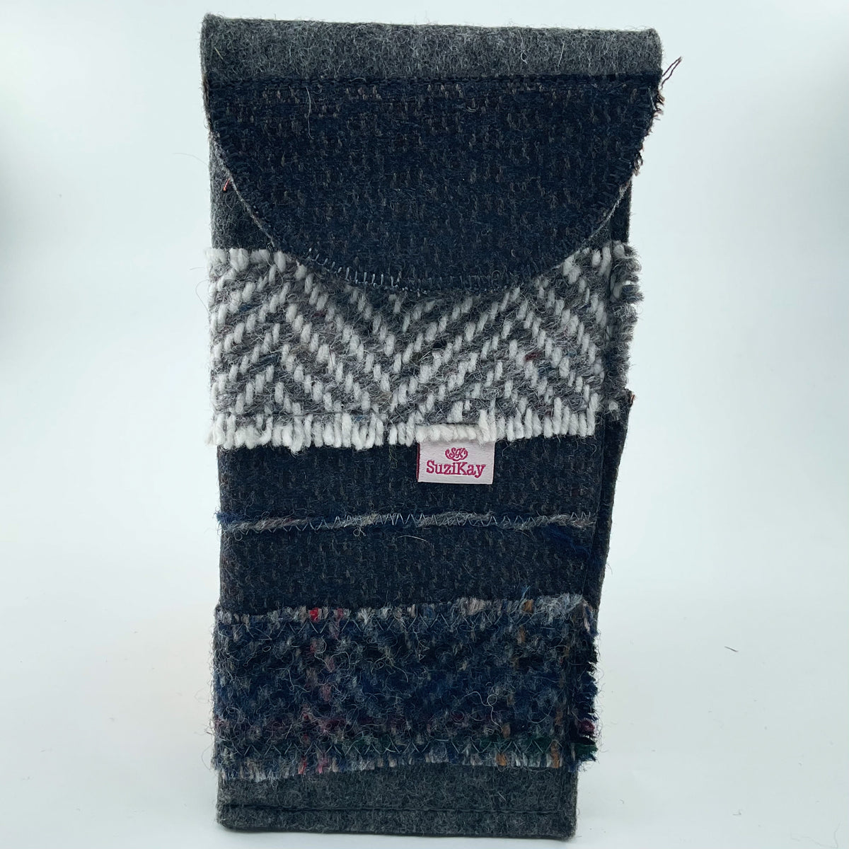 A handmade fabric pouch or sleeve made from layered wool and tweed fabrics in dark navy, charcoal gray, and white, featuring a small pink and white Suzikay brand label.
