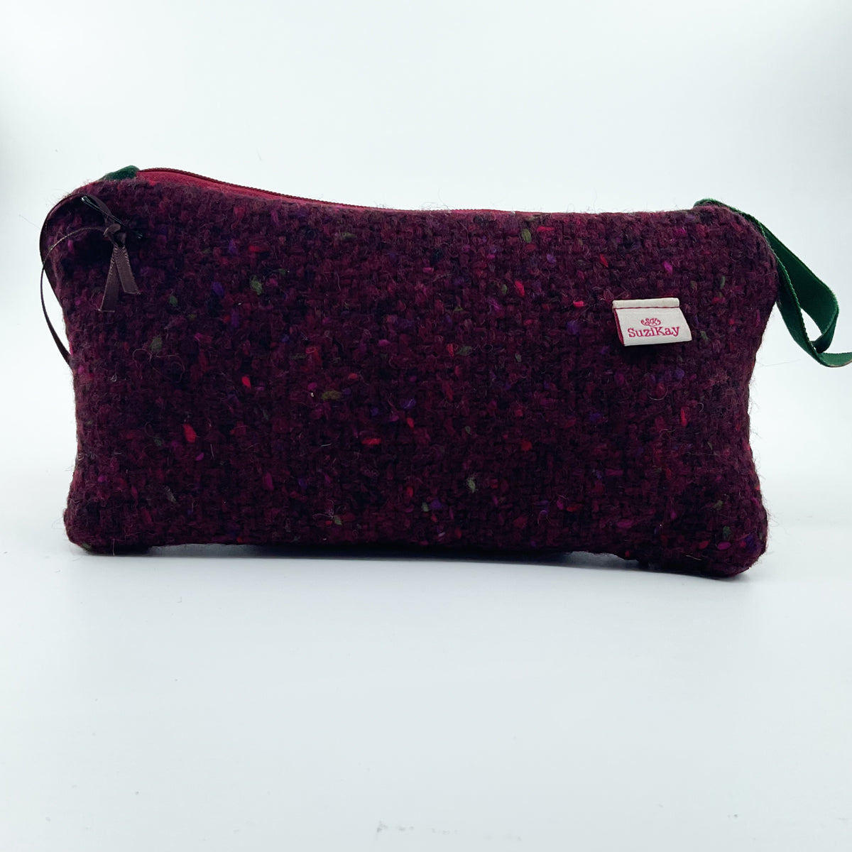 A textured tweed cosmetic pouch in deep burgundy with multicolored flecks, featuring a small white SuziKay brand tag and a green strap on a white background.