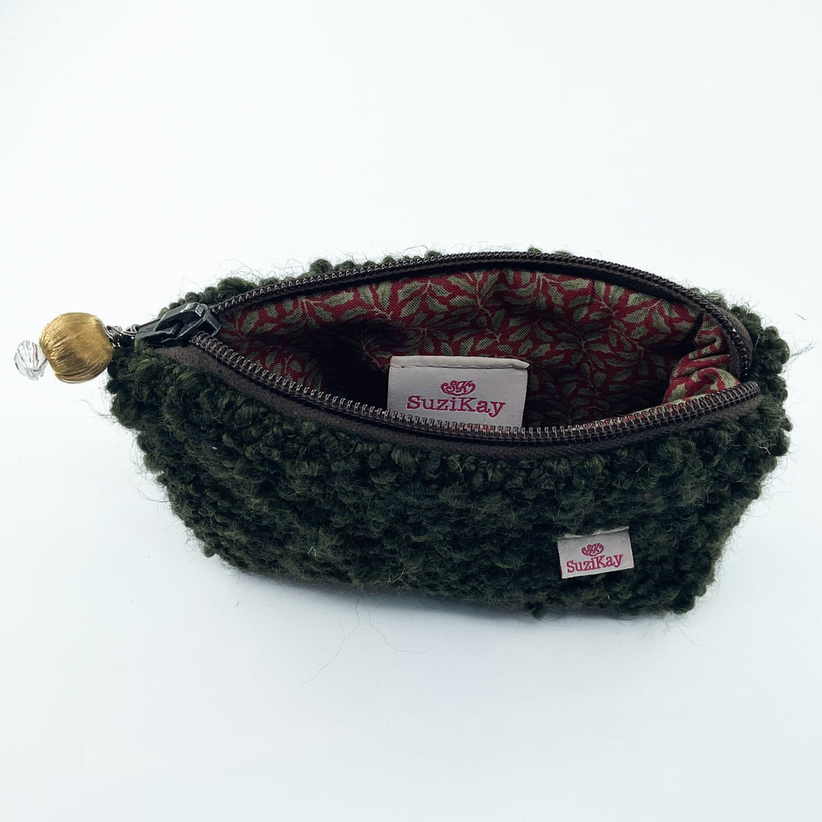 An olive green textured faux fur zippered pouch with a patterned red and green lining, featuring a wooden bead charm on the zipper pull and a visible Suzikay brand tag.