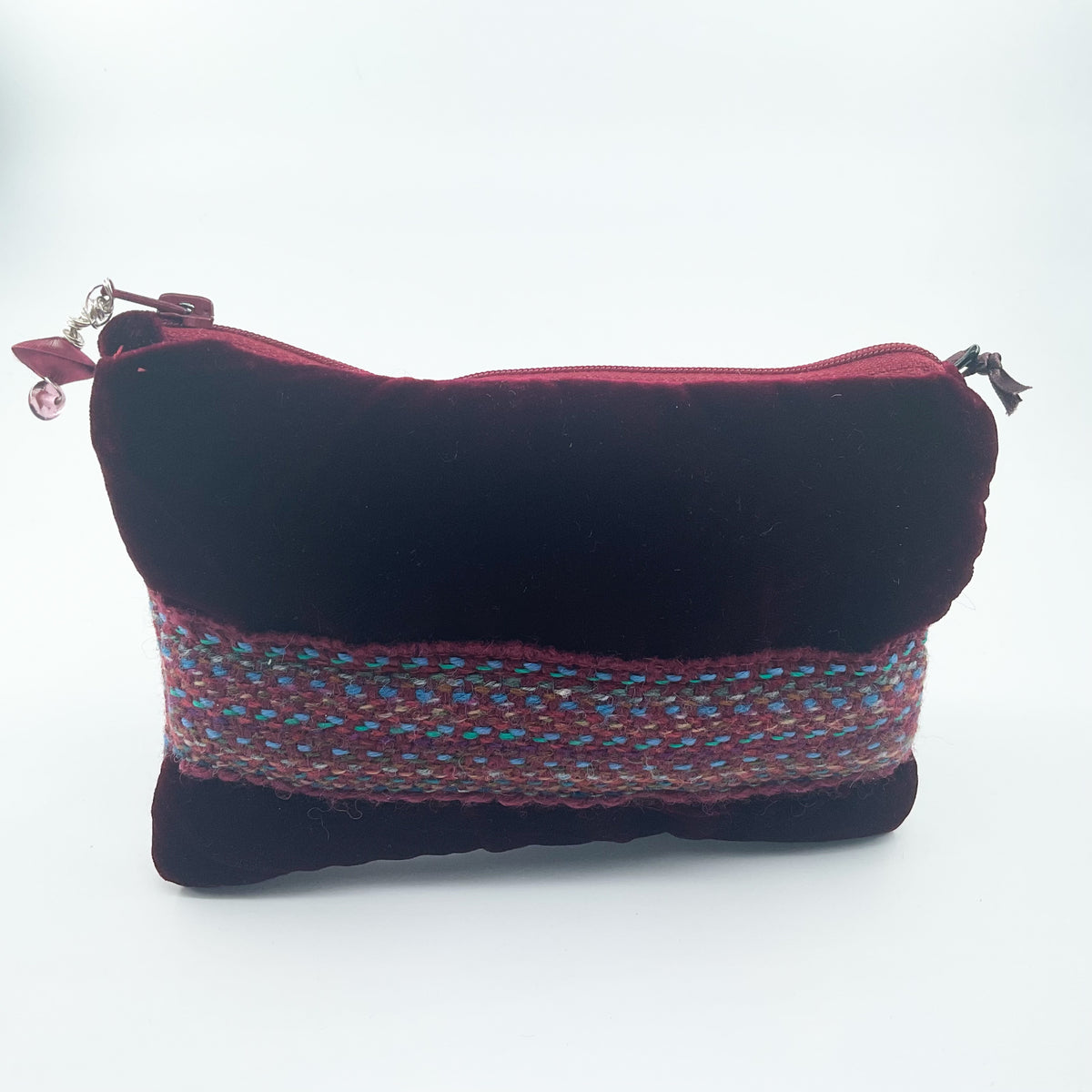 A small rectangular velvet pouch in deep burgundy with a wide woven multicolor band across the middle and a silver zipper closure, shown on a plain white background.