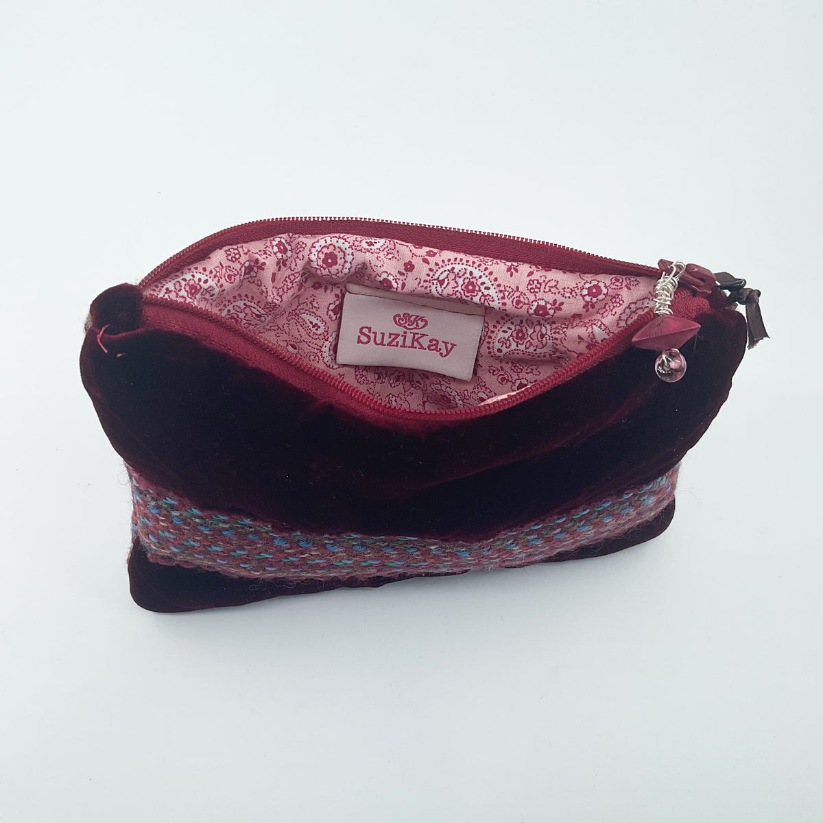 A dark burgundy velvet cosmetic pouch with a patterned pink lining and a woven multicolor band near the bottom. The brand name 'Suzikay' is visible on the interior label.