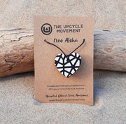 A necklace with a heart-shaped pendant featuring a black and white geometric pattern, displayed on a wooden surface with a brand tag attached.