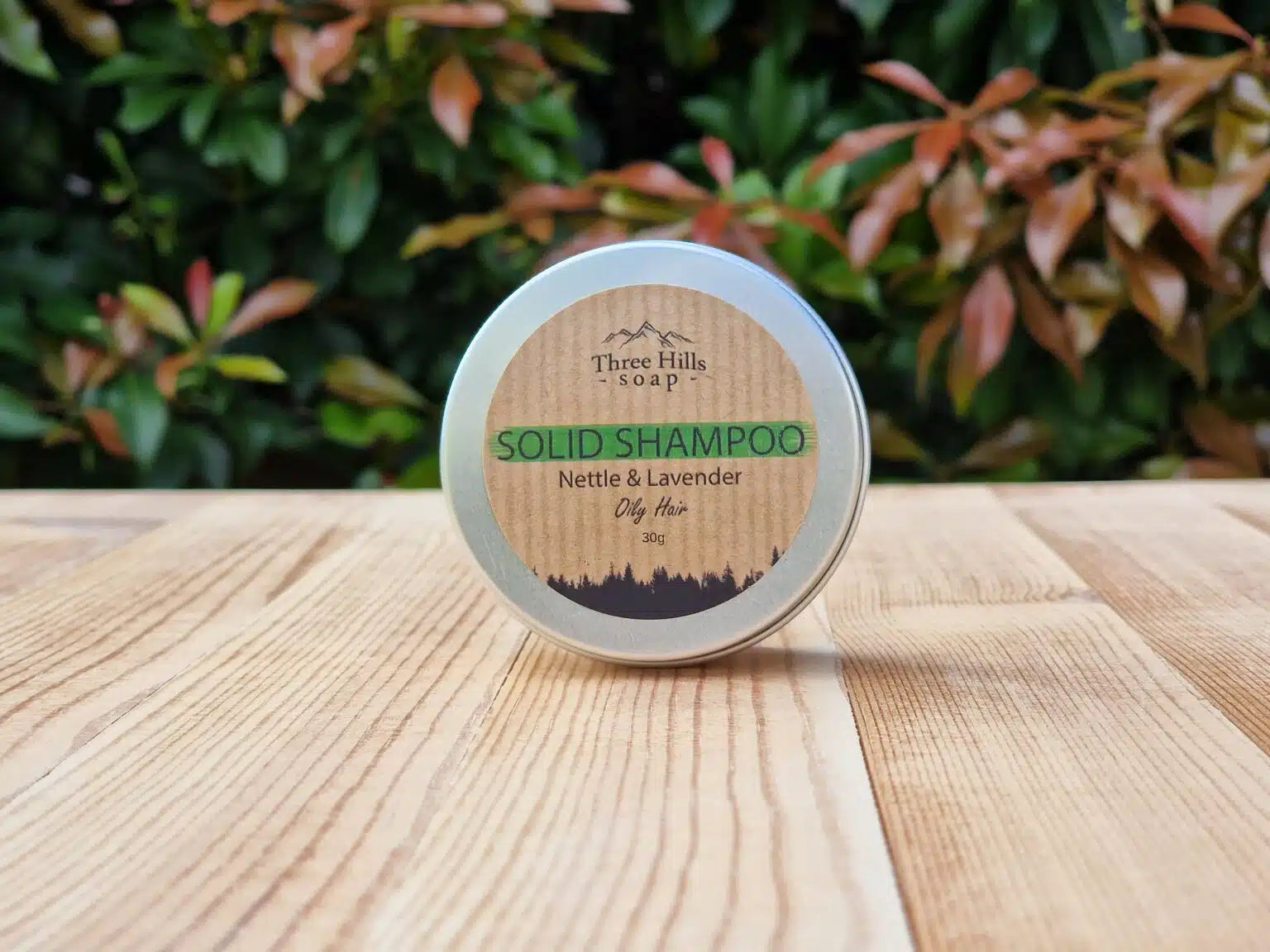 A round tin of Three Hills Soap Solid Shampoo (Nettle & Lavender) sits on a light wooden table with a blurred green and reddish foliage background.