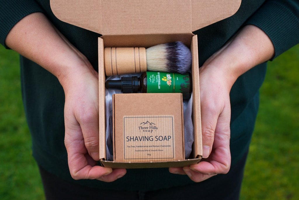 Two hands holding a cardboard box with shaving products including a brush, soap, and other items.