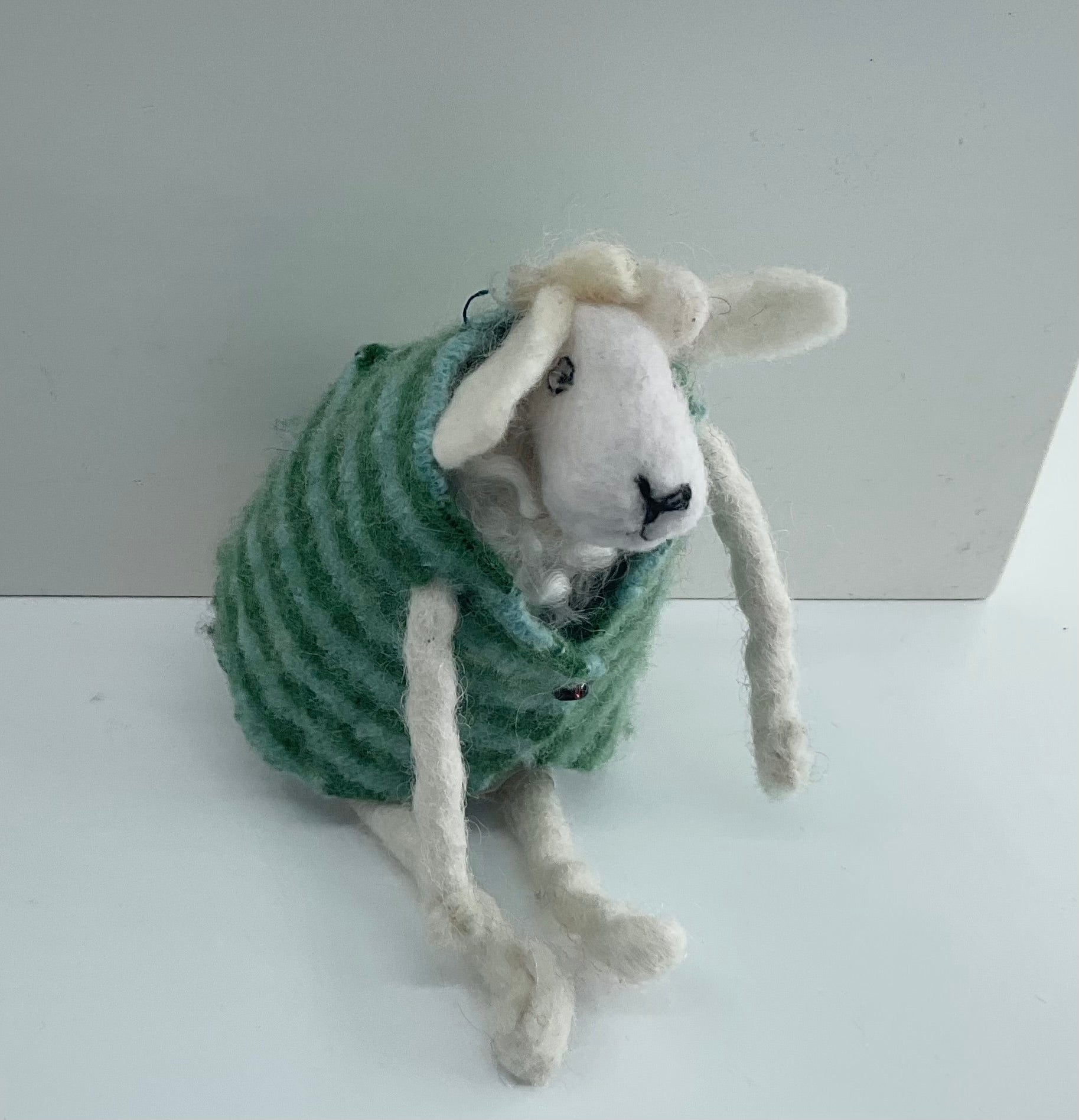 Felted sheep wearing a green sweater on a white surface with a light gray background
