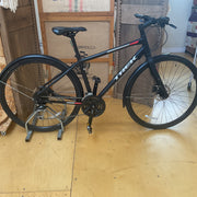 A black Trek bicycle with a rear rack and fenders, displayed on a metal stand in a workshop or garage setting with cardboard boxes in the background.