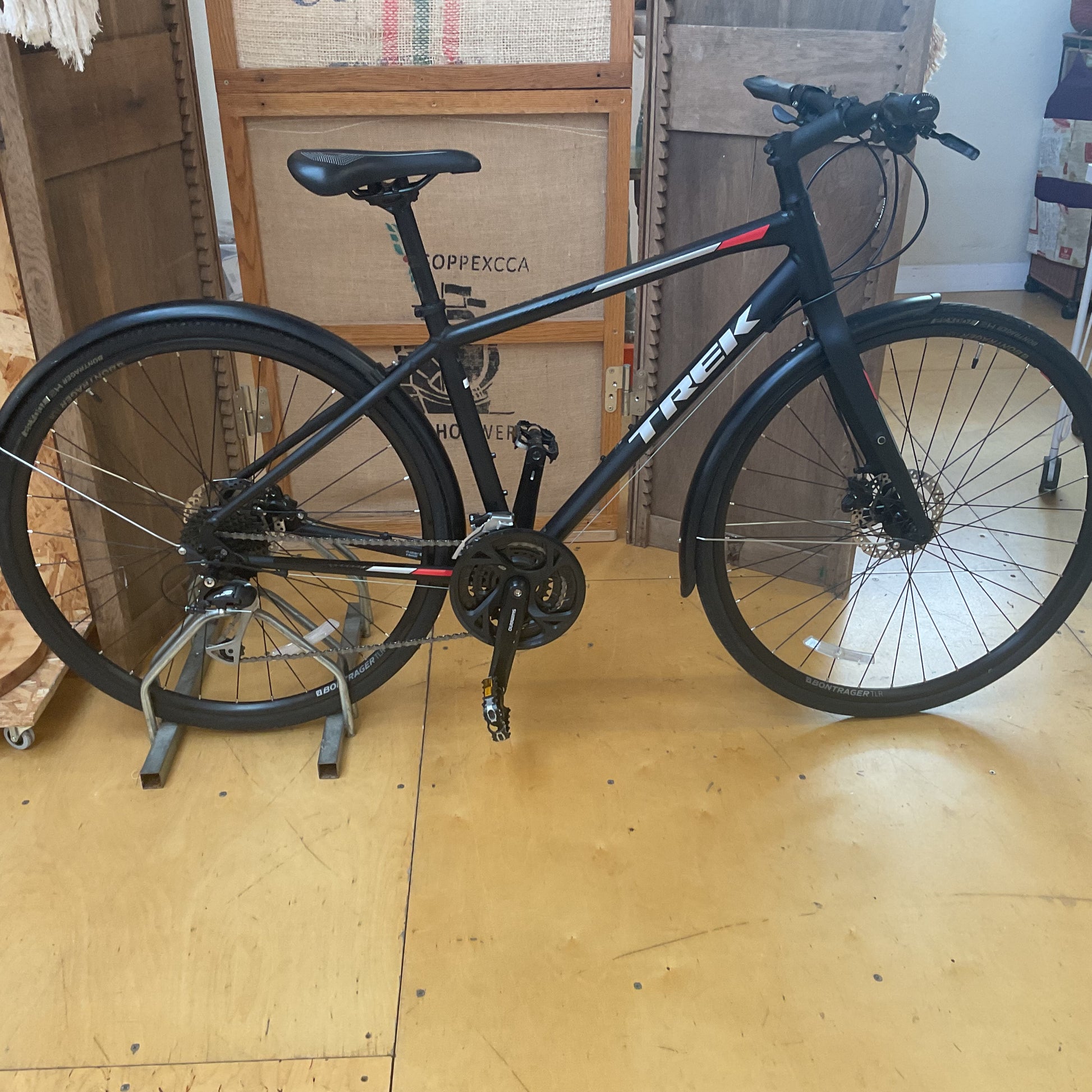 A black Trek bicycle with a rear rack and fenders, displayed on a metal stand in a workshop or garage setting with cardboard boxes in the background.