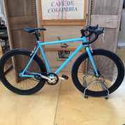 A bright turquoise road bicycle with black components, displayed on a metal stand in front of wooden coffee roaster crates.