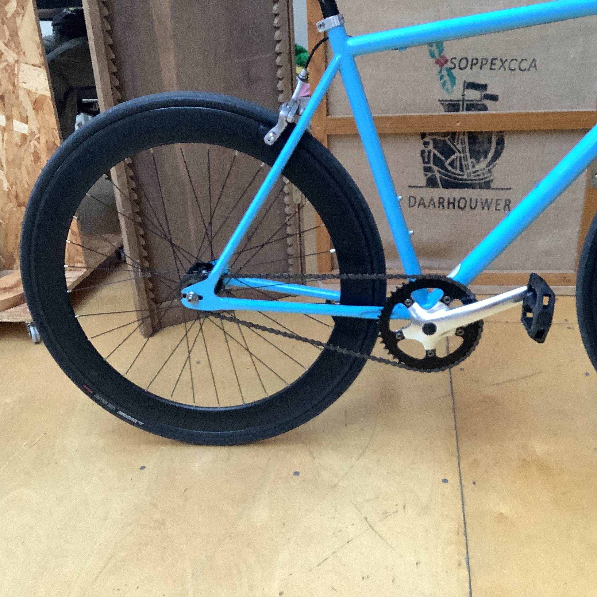 A close-up of a bright blue bicycle frame and rear wheel with a black tire, shown indoors on a wooden floor in front of a cardboard box.