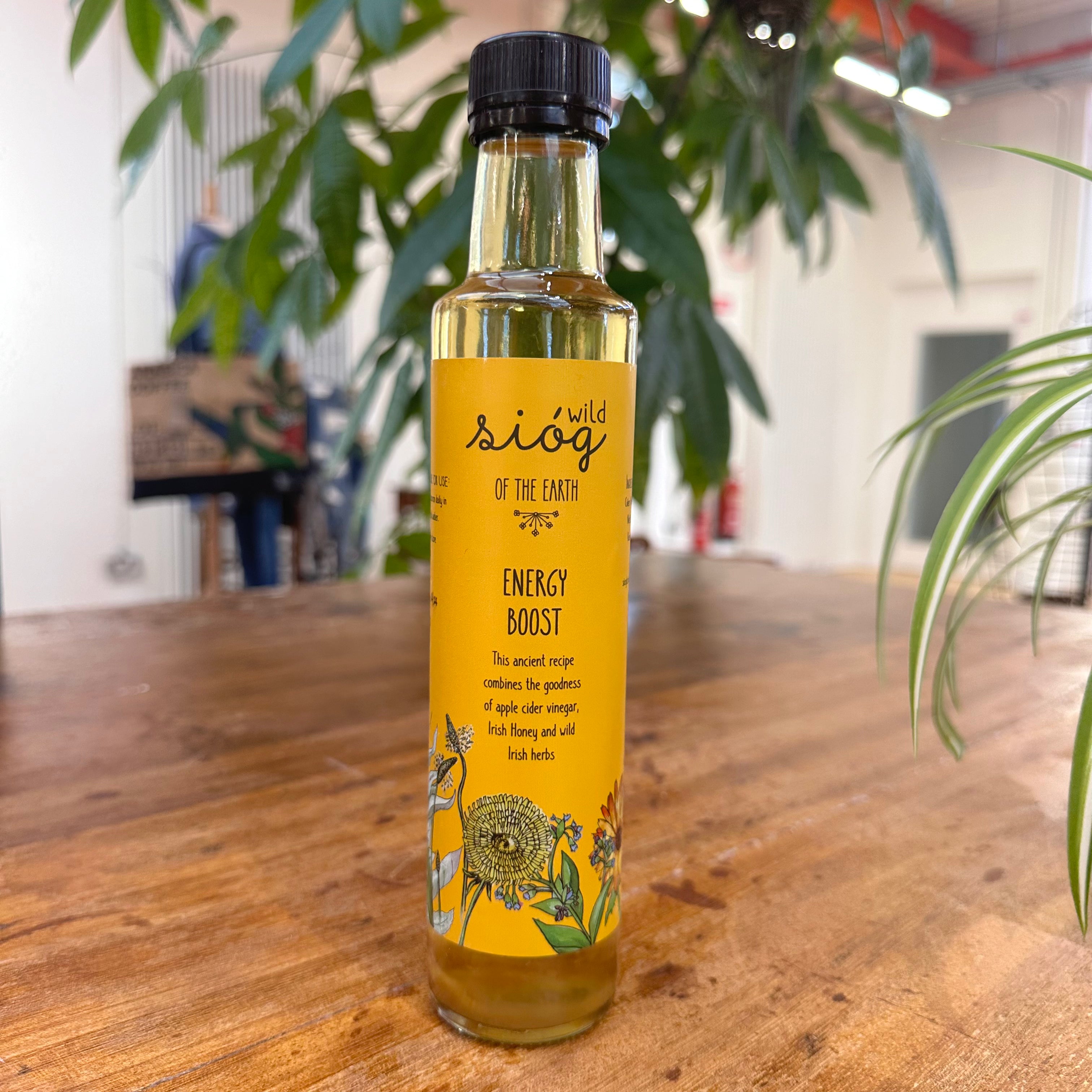 A bottle of Sióg of the Earth Energy Boost Tonic placed on a wooden surface with plants in the background.