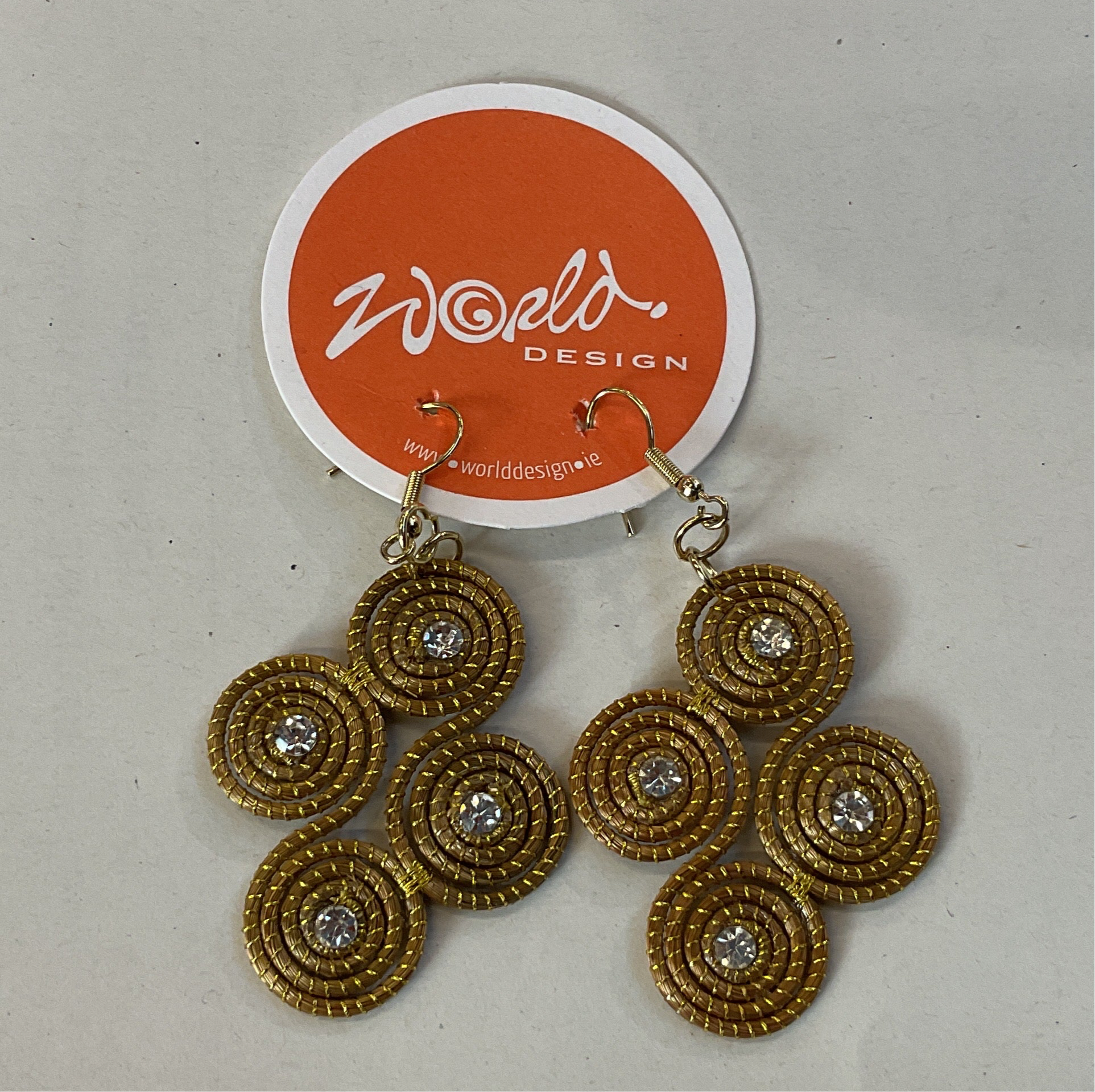 A pair of gold-colored capim dourado grass earrings with circular patterns and silver accents, displayed on a red and white card with 'World Design' branding.
