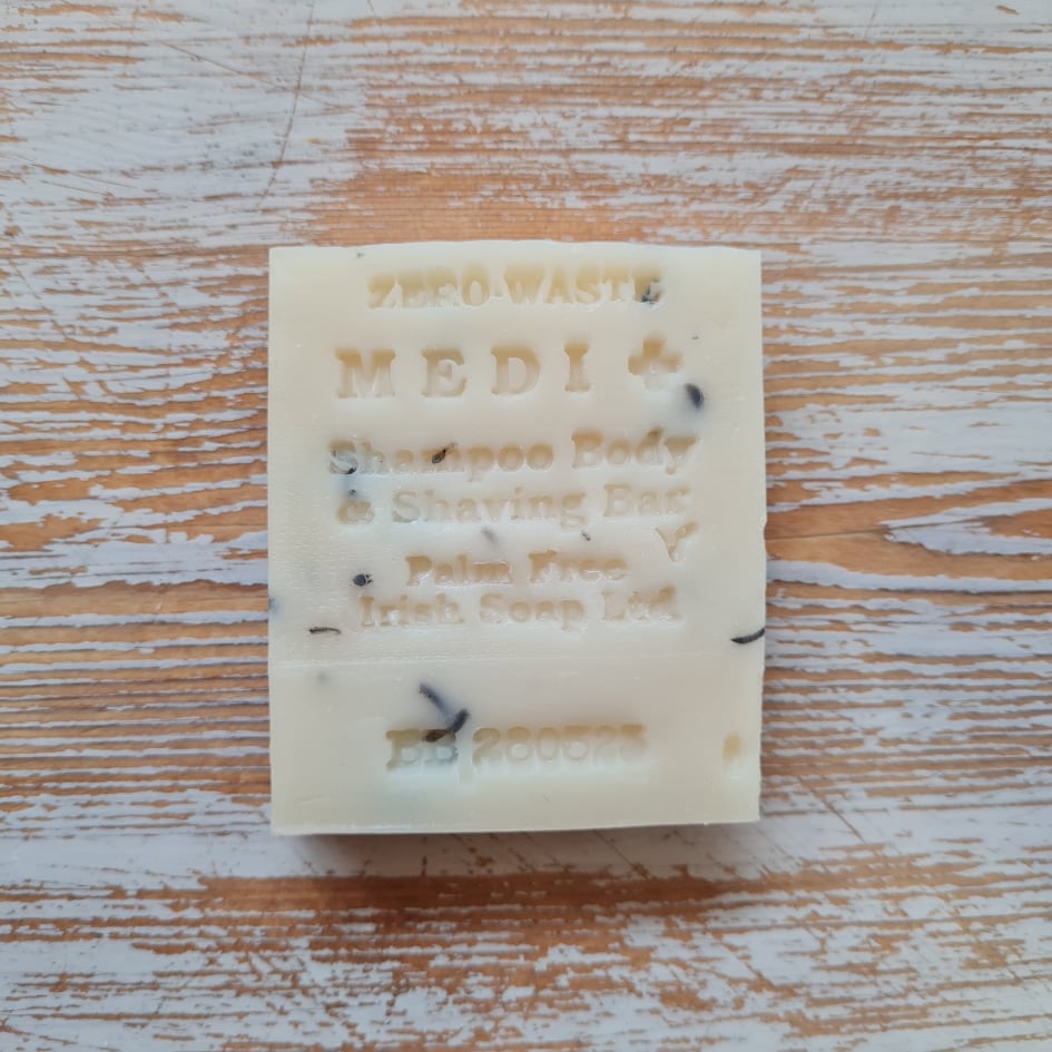 A white shampoo bar with 'MEDI' branding and various product labels including 'Shampoo Body & Shaving Bar' and 'Irish Companies Ltd'. The bar is placed on a wooden surface with visible cracks.