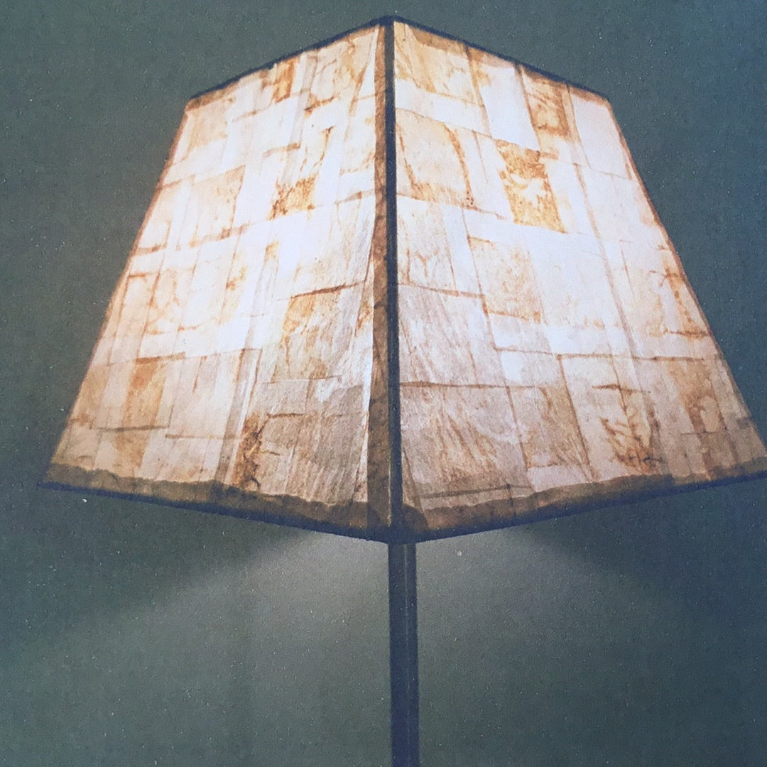 A lamp shade made out of paper tea bags with a beige and brown color, featuring a unique geometric shape and placed on a stand.